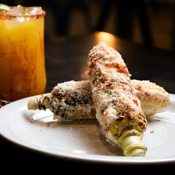 Two elotes, grilled corn on the cob with cheese and chili powder, on a white plate with a drink.