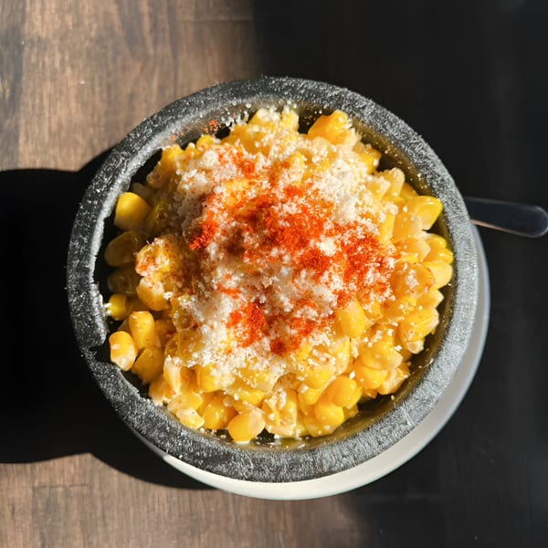 Elote in a dark bowl, topped with white cheese and red chili powder, on a wooden table.