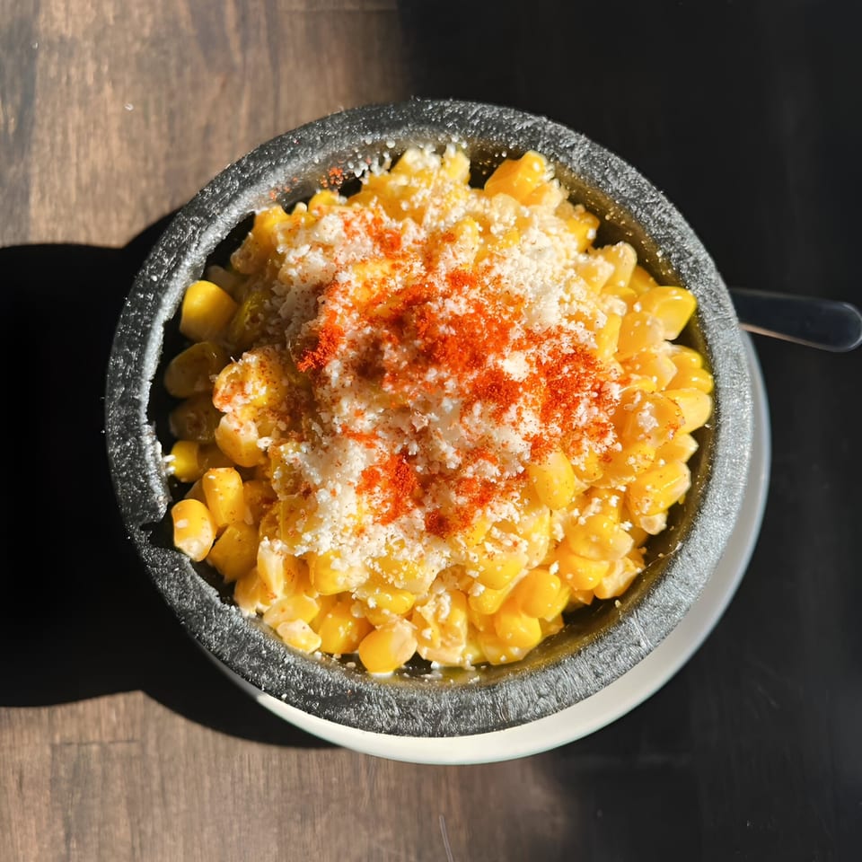 Elote corn in a dark bowl, topped with white cheese and red chili powder, on a wooden table.