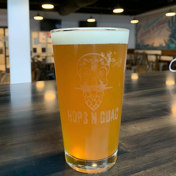 A Hops N Guac branded pint glass filled with a light amber beer and foamy head.