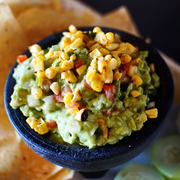 A black bowl of Hops N Guac's guacamole topped with corn, tomatoes, and onions, with tortilla chips.