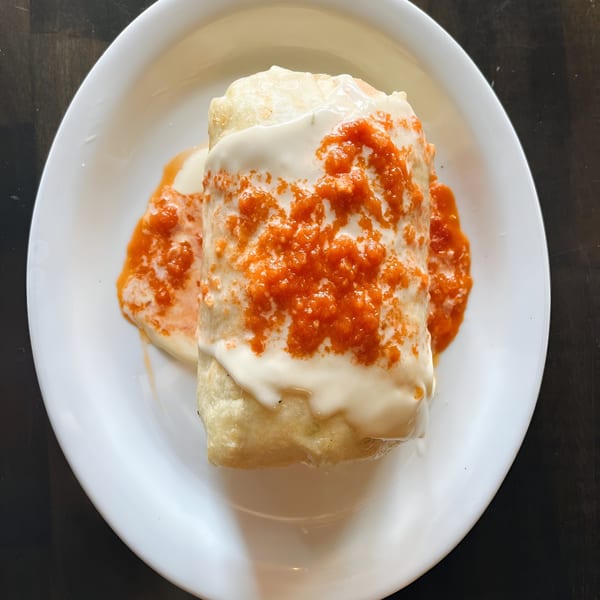 A Hops N Guac burrito covered in white sauce and red salsa on a white plate.