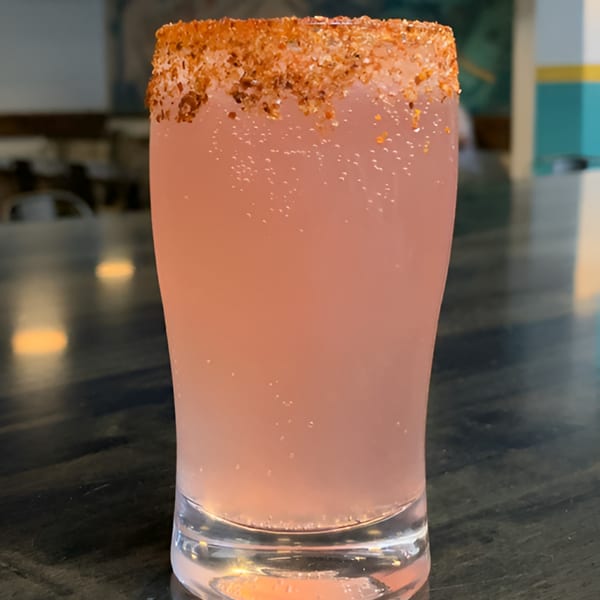 A pink, bubbly drink with a chili-salt rim sits on a dark table at Hops N Guac.