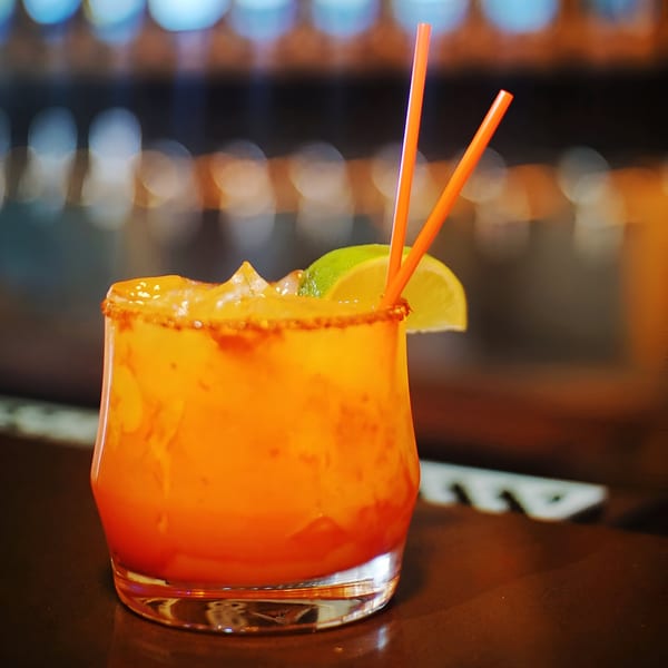 A vibrant orange margarita with a chili-lime rim, lime wedge, and two orange straws on a bar.